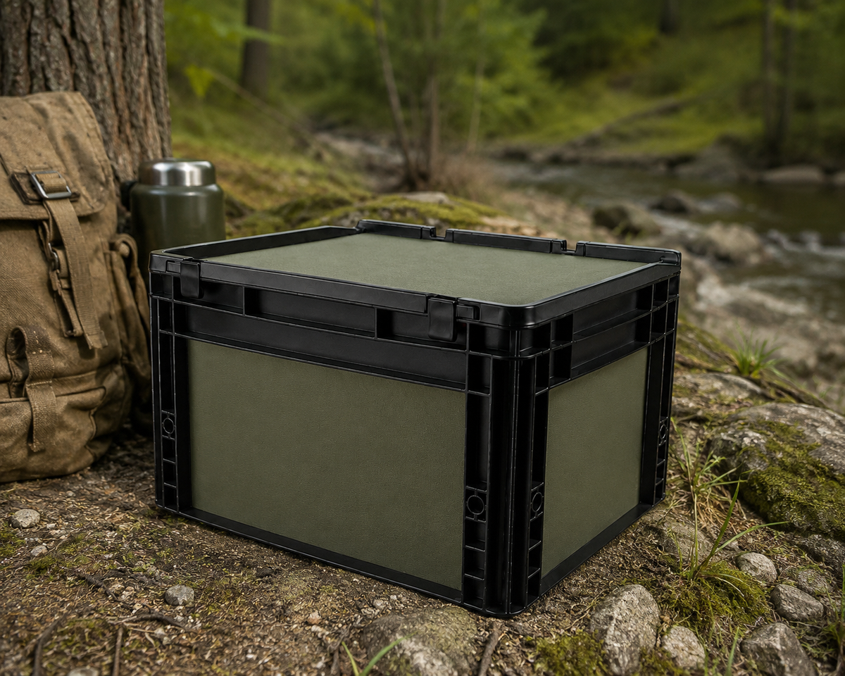 OctoVan Eurobox in matt Olivegrün mit Scharnierdeckel auf unebenem Waldboden zwischen Steinen, Wurzeln und Moos. Neben der Box stehen ein Outdoor-Rucksack und eine Thermoskanne. Die Szene vermittelt Vanlife-, Camping- und Outdoor-Abenteuer in natürlicher Waldumgebung.