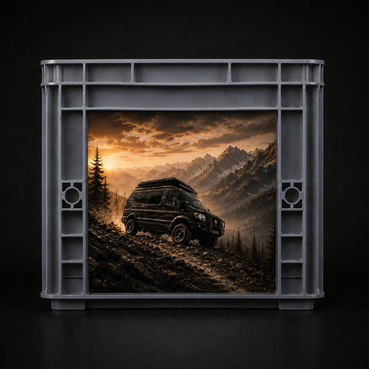 Grauer Kunststoffrahmen mit rechteckiger Öffnung, darin ein Foto eines schwarzen Offroad‑Vans auf steinigem Berghang bei Sonnenuntergang. Im Hintergrund sind Tannen, felsige Gipfel und goldene Wolken zu sehen.